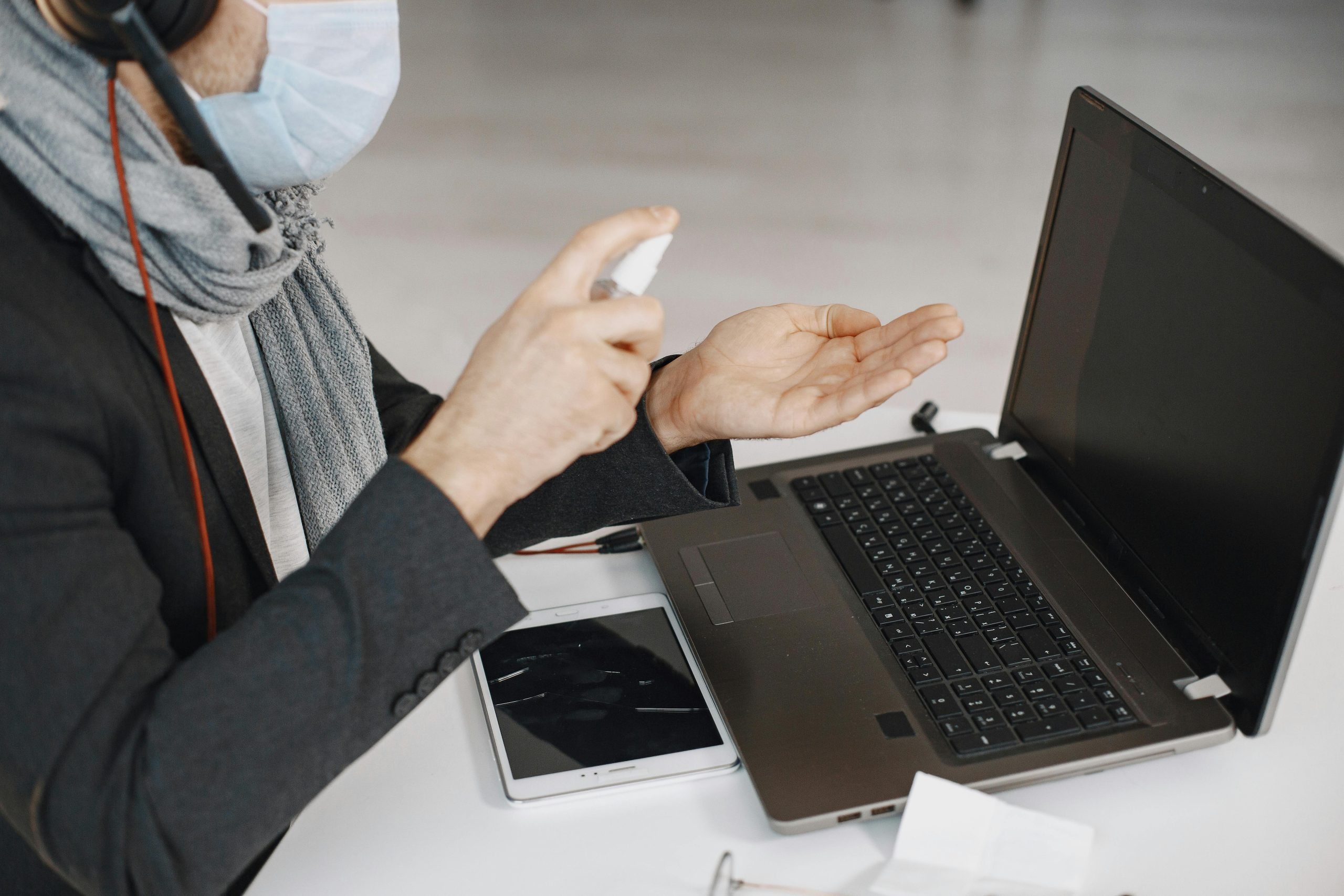 A man wearing a mask sanitizing his hands while working at a laptop indoors.
